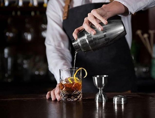 A bartender pouring a mixed drink into a glass with a lemon rind