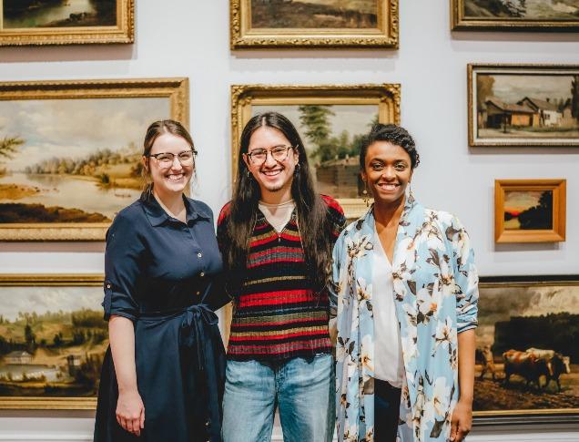 Three people stand in front of a wall of paintings at Museum London.