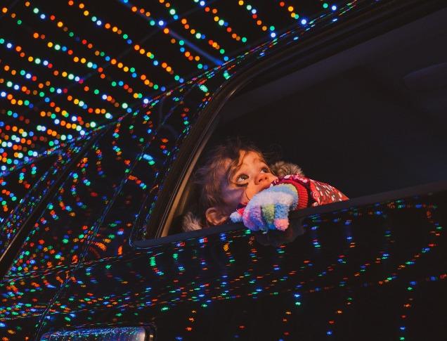 A kid looks out of a car window as the vehicle passes through a magical tunnel of holiday lights.