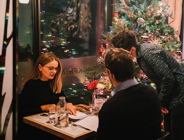 A couple dining at a restaurant with a server looking on as they order food with a Christmas tree behind them