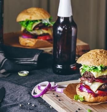 Two gourmet burgers resting on a wooden plate with two beers on the side