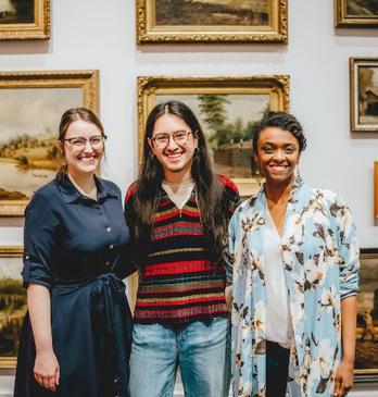 Three people stand in front of a wall of paintings at Museum London.