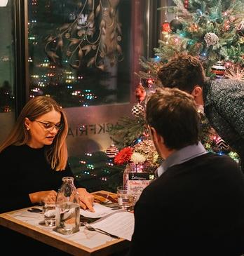 A couple dining at a restaurant with a server looking on as they order food with a Christmas tree behind them