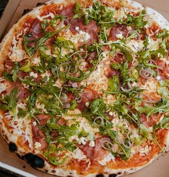 aerial view of a pizza on a black marble table loaded with pepperoni, sausage and basil and a basket of chicken wings