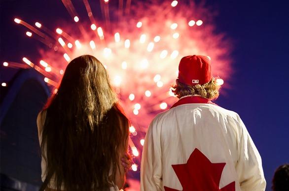 Two people viewed from behind looking at red fireworks lighting up the sky, one individual has a Canada flag jacket