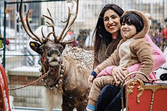 A mother and her child seated on a red sleigh with a deer behind them