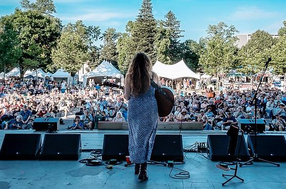 A female playing her guitar on stage with a large crowd gathered outdoors at the Home County Music & Art Festival held in London, Ontario.