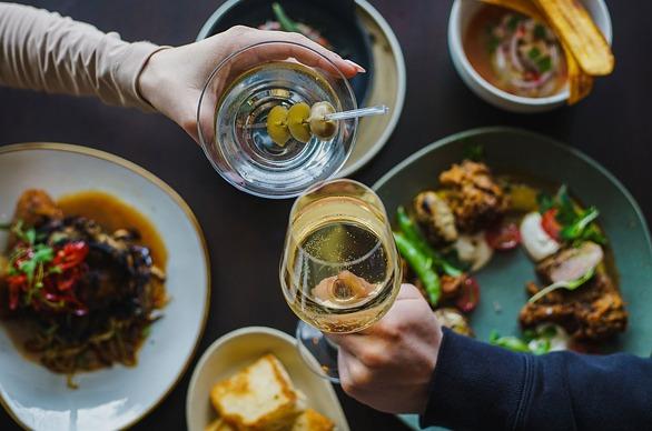 A couple holding their beverages over a table with various plated dishes at a restaurant.