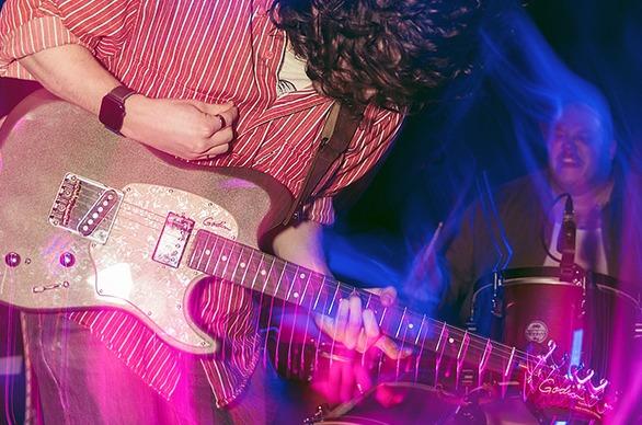 A band playing live on stage, the focus is on a musician playing the electric guitar and in the background is a person playing drums