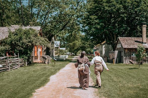 Two females walking down a dirt path by old wooden homes from the 1800s in Fanshawe Pioneer Village, located in London, ON