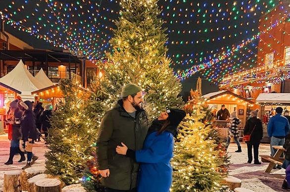 A couple hugging each other in front of a large, bright decorated Christmas Tree at the Merry Market held at 100 Kellogg Lane