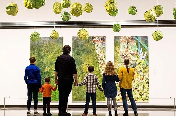 A family all holding hands, viewed from behind, looking at paintings on display at Museum London, located in London, Ontario