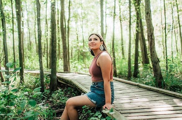 A female siting on the edge if a wooden path surrounded by tall trees in Sifton Bog, located in London, Ontario