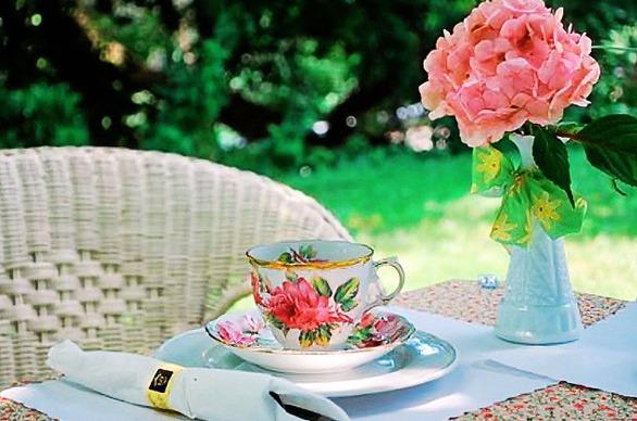 A tea cup set up at a table with a pink flower in a vase in an outdoor setting.