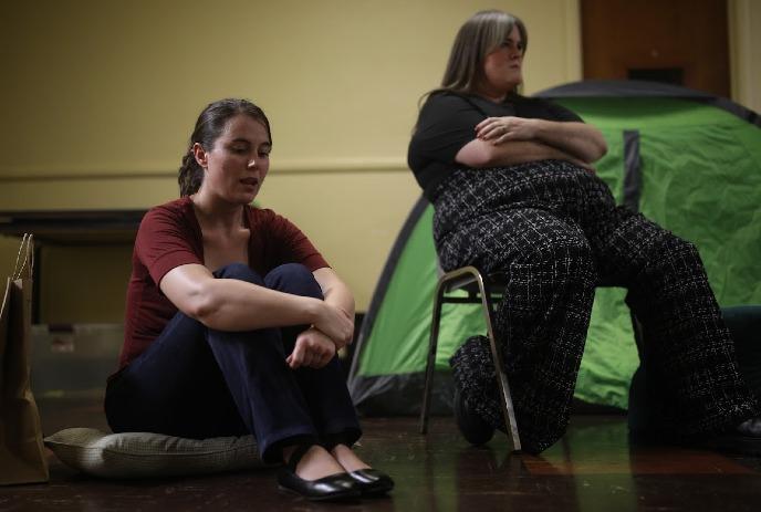 Two people sit indoors near a green tent, resting on chairs and cushions in a shelter setting.