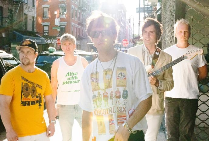 A group photo of the band The Brokes standing on a city street with one member holding a guitar.
