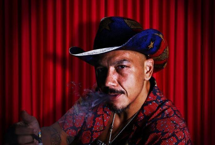Luis J Gomez with a mustache in a red patterned shirt and a star-spangled cowboy hat smokes against a vibrant red curtain.