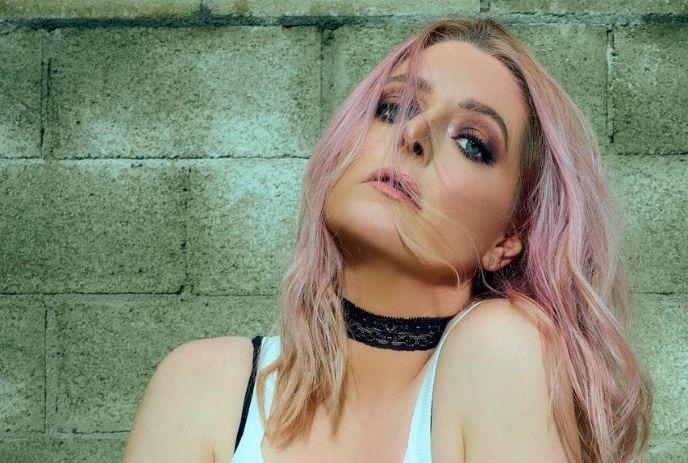A photo of Lauren Ash with pink hair, wearing a white top and black lace choker, posing against a textured green wall.