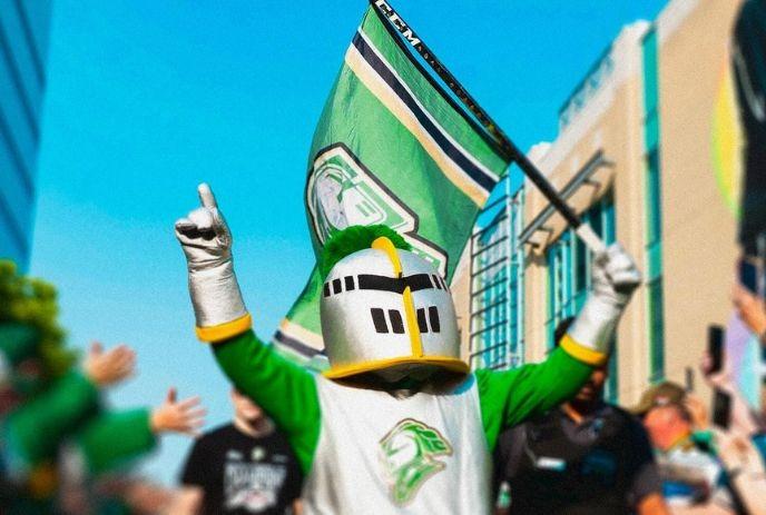 The London Knight holding a London Knights flag in one hand, and pointing to the sky in the other, outside in a crowd.