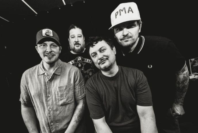 Four men of Illscarlett are standing together in a black and white image. Two of them are wearing caps.