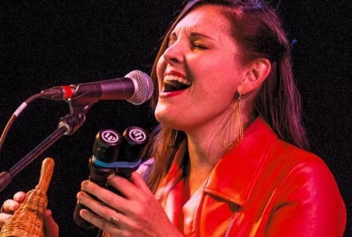 A woman wearing a red jacket sings passionately on stage into a mic.