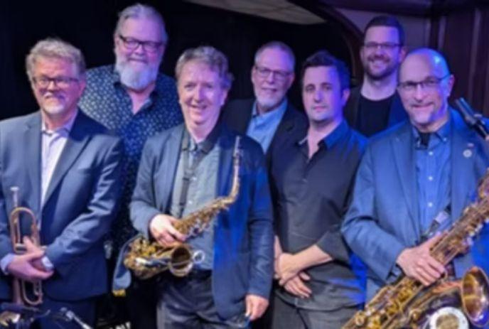 A group of seven men dressed in suits, smiling on stage with musical instruments.