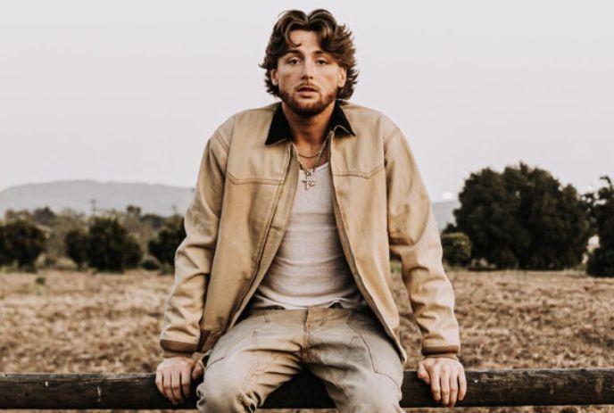 Bailey Zimmerman sits on a fence in a dry field with mountains and trees, wearing a tan jacket.