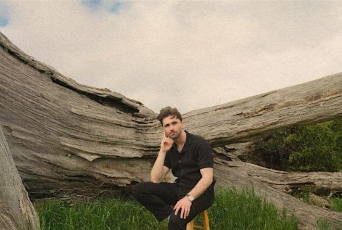 John Muirhead sitting on a stool outside in front of a large tree trunk on a cloudy day.
