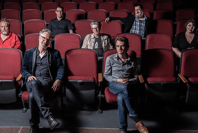 Seven artists seated in red theater rows with focus on front pair in dim cinematic setting.