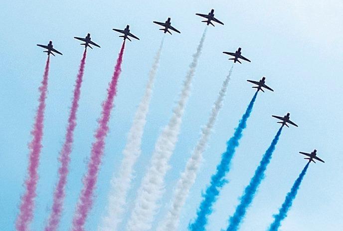 Nine aircraft in diagonal formation, trailing colourful smoke in clear blue sky, performing synchronized aerobatic maneuver.