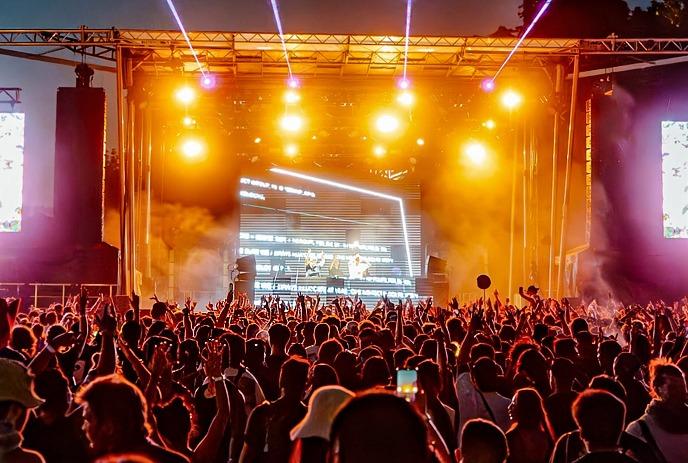 A bright lit night concert with DJ on stage, crowd cheering under beams of light and glowing screens.