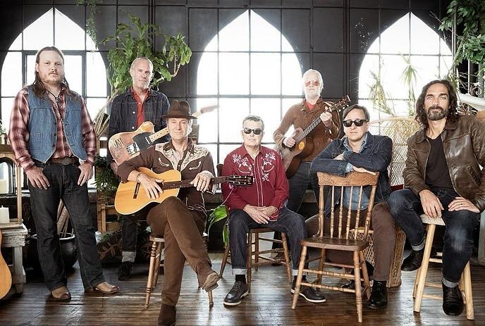 Six band members of Blue Rodeo standing outdoors in front of a modern building with large windows, posing for a picture.