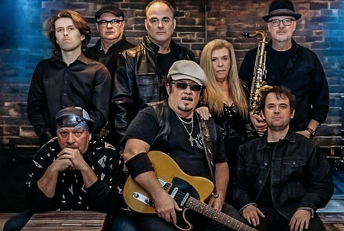 Eight artists pose together with hats sunglasses and saxophone against neutral gray backdrop.