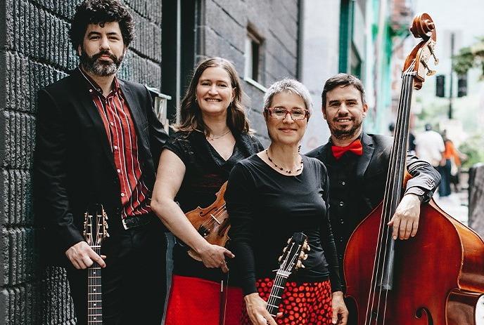 Four musicians pose with instruments outdoors by windows, smiling for a picture.