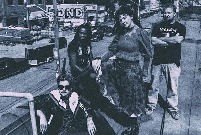 A group of four people stand and sit on a rooftop, overlooking a cityscape with street signs and buildings.