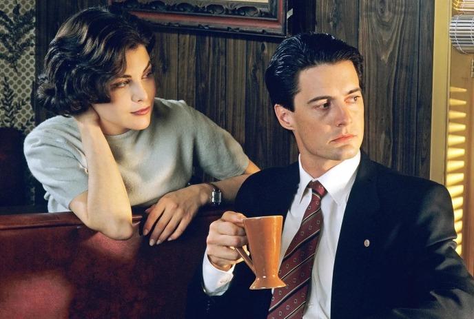 Two people sit in retro diner booth, one holding mug while the other leans over.