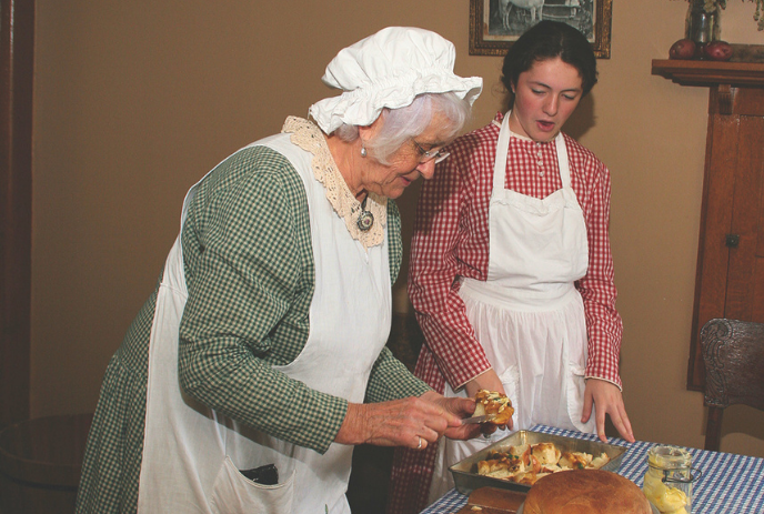 Two women in old style dresses serving food on table set with bread butter and baked dish.
