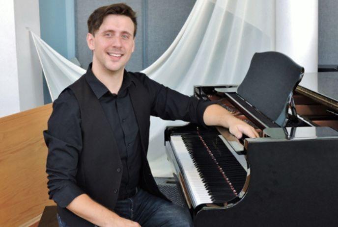 Alex Baerg in black attire poses at grand piano in studio with white drapes and wood panel backdrop.