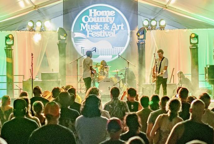 A band performs on a lit stage under a large tent at a music festival with crowd cheering.