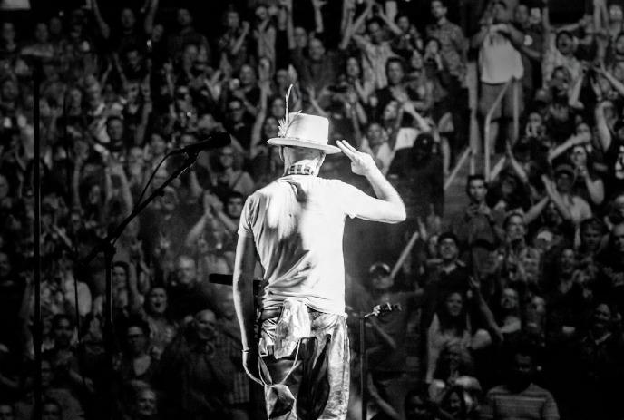 In a black-and-white image, a performer in a hat salutes an applauding, packed stadium crowd.