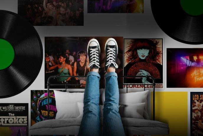 Feet in sneakers are propped up on a bed, surrounded by wall posters of musicians and large vinyl records.
