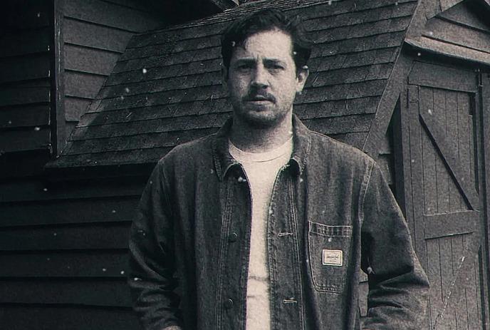 Black and white portrait, artist in denim jacket stands before rustic shed, hands in pockets, posing for a picture.