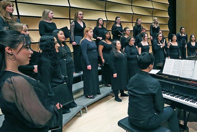 Choir in black outfits as pianist plays, set against acoustic wall in concert hall setting.
