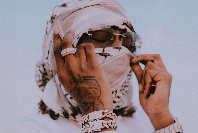 Northsidebenji in white, scarf on, holding a cigarette near his tattooed hand with jewelry, against a clear sky.