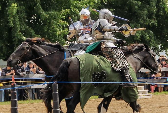 Two armored riders on horses meet in a friendly gesture inside an indoor arena setting.
