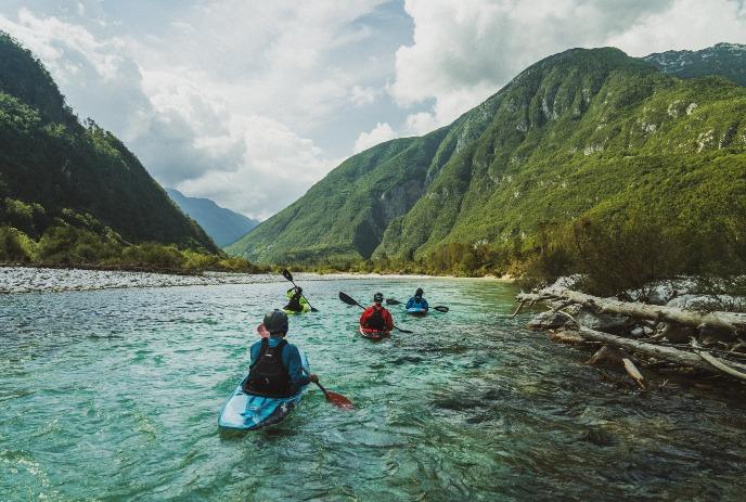 Alt text: A group of kayakers navigating a clear mountain river surrounded by greenery and steep hills under a cloudy sky.