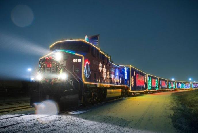 Holiday train glows with festive lights, peace sign, Santa, and trees; snow-covered track at night.