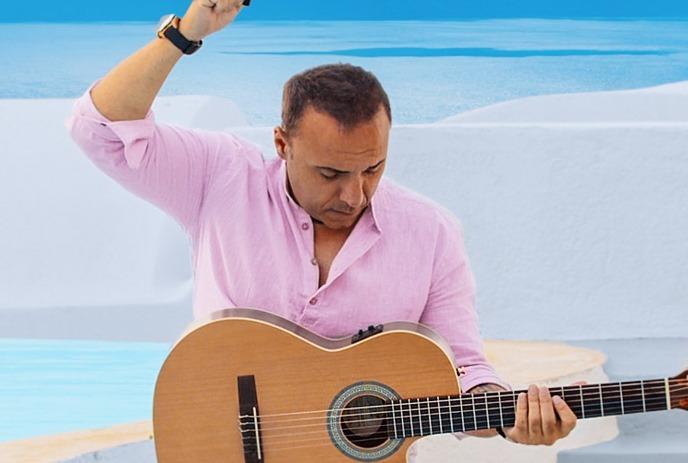 Artist in bright pink?shirted person sitting outdoors by white Mediterranean architecture, holding a guitar with blue sea .