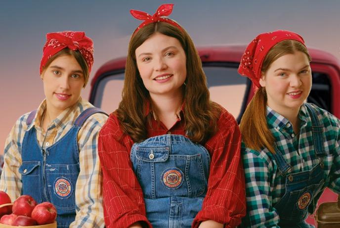 Three women in red bandanas and overalls pose in front of a truck with apples.