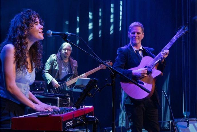 Three musicians performing on stage with guitars and keyboard under blue and purple lighting.